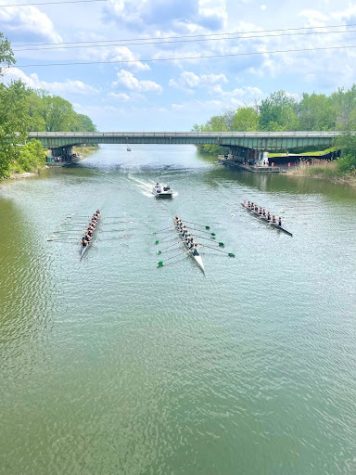 A distant shot shows several boats competing in competition passing under a bridge.