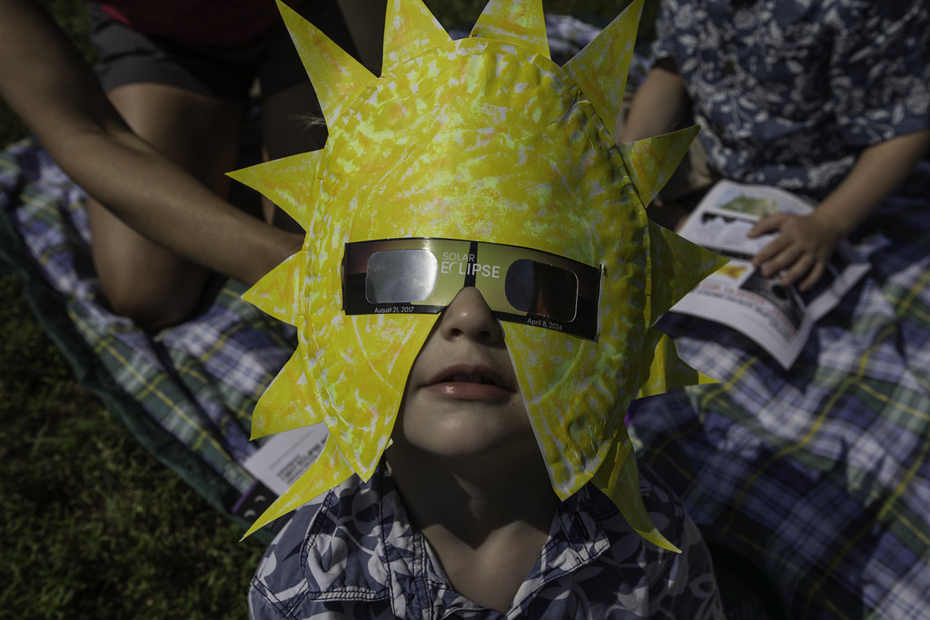 A child wears the appropriate glasses that are surrounded by a yellow paper sun mask.