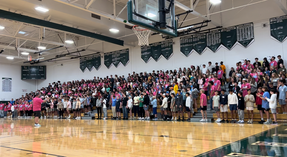 First year students dressed in pink stand in bleachers while the principal speaks to them from the center of the floor.