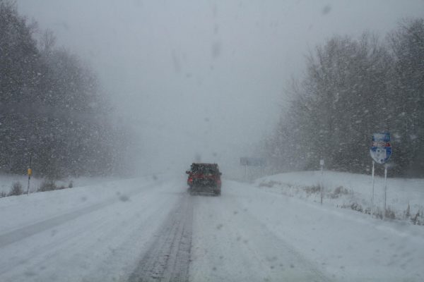 A shot from behind an orange vehicle braking on the highway as snow falls and the roads are covered with snow.