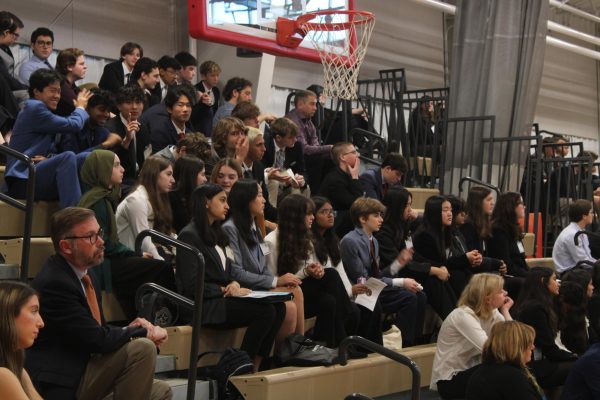 Students in professional dress sit in the gym bleachers, listening to a speaker.