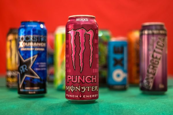 Colorful energy drinks sit on a green table cloth. The feature drink is Monster Punch in a pink can. The cans are in front of a red background.