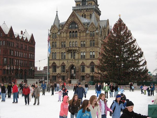 Ice skaters make their way around the rink. The famous tree is all lit up with colored lights in the background.