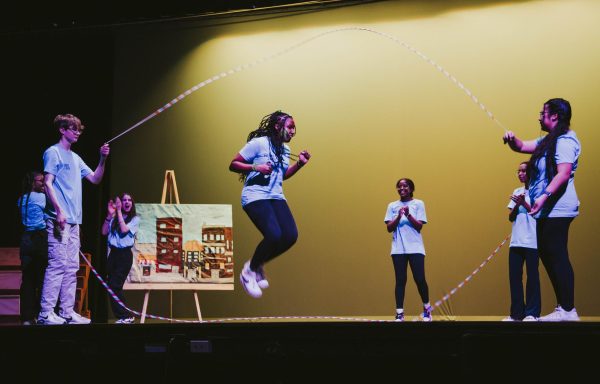 A performer demonstrates double dutch jump roping technique while other performers cheer her on.