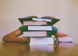 A student hides behind a pile of books sitting at a table. Arms and shoulders are visible, but not a head or face.