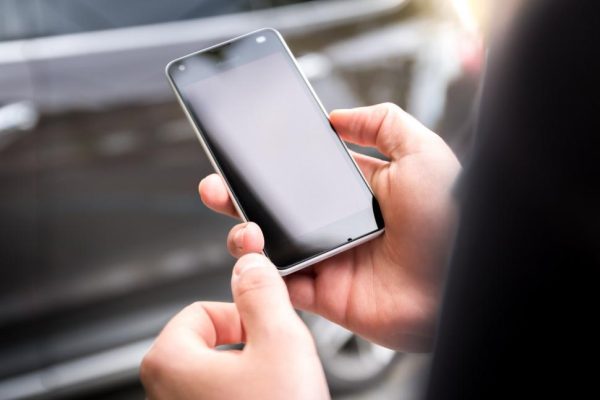 A person is seen holding a cell phone in their hand, with the device visible and prominently displayed. The individuals fingers are gripping the phone, and the screen is likely illuminated.