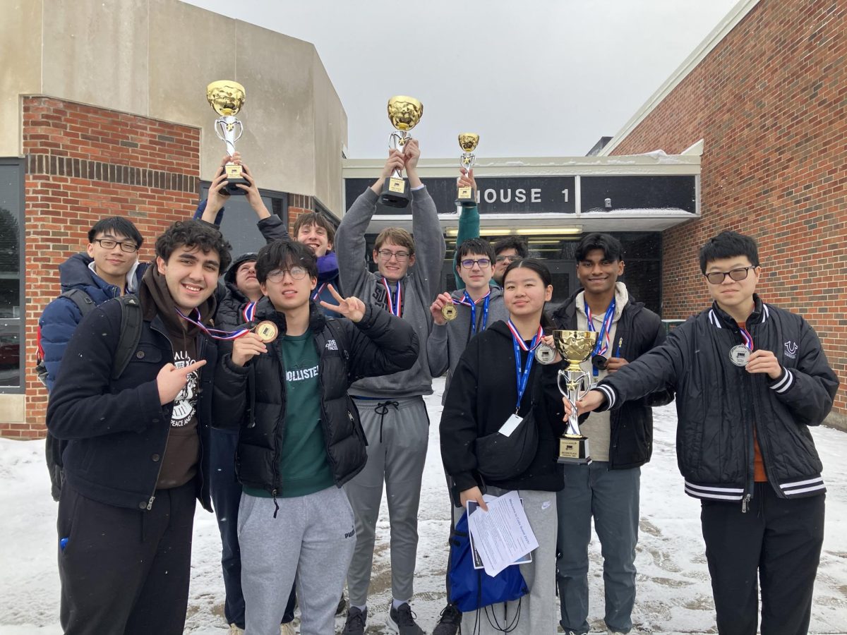 Students stand in front of the high school with trophies raised up over their heads.