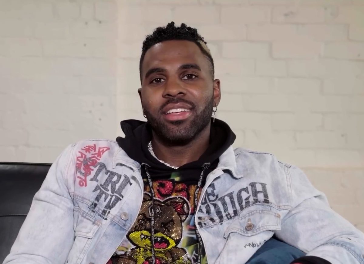 Derulo is sitting on a black leather couch for the interview. He is wearing a black hoodie, layered with a sequined t-shirt that has a colorful angry bear on it. He has a faded jean jacket on over the top.