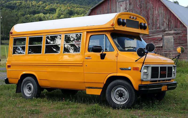 An older half-sized school bus sits in front of a barn. The bus is still yellow but has been stripped of all lettering, including school affiliation.