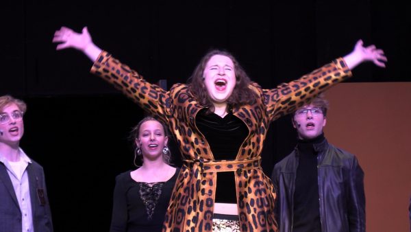 A lead singer has her eyes closed, hands in the air, wearing a leopard style jacket.