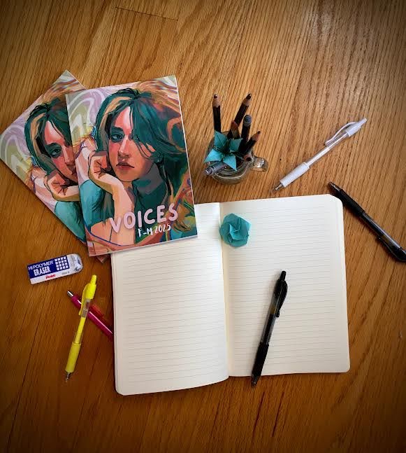 A desk table displays an open, blank notebook with copies of the publication and various writing utensils surrounding it.