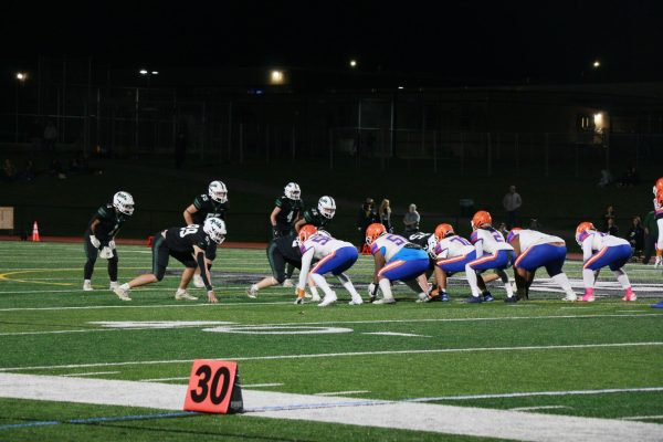 The visiting team in white/blue/orange hikes the ball as the green home team moves to stop them.