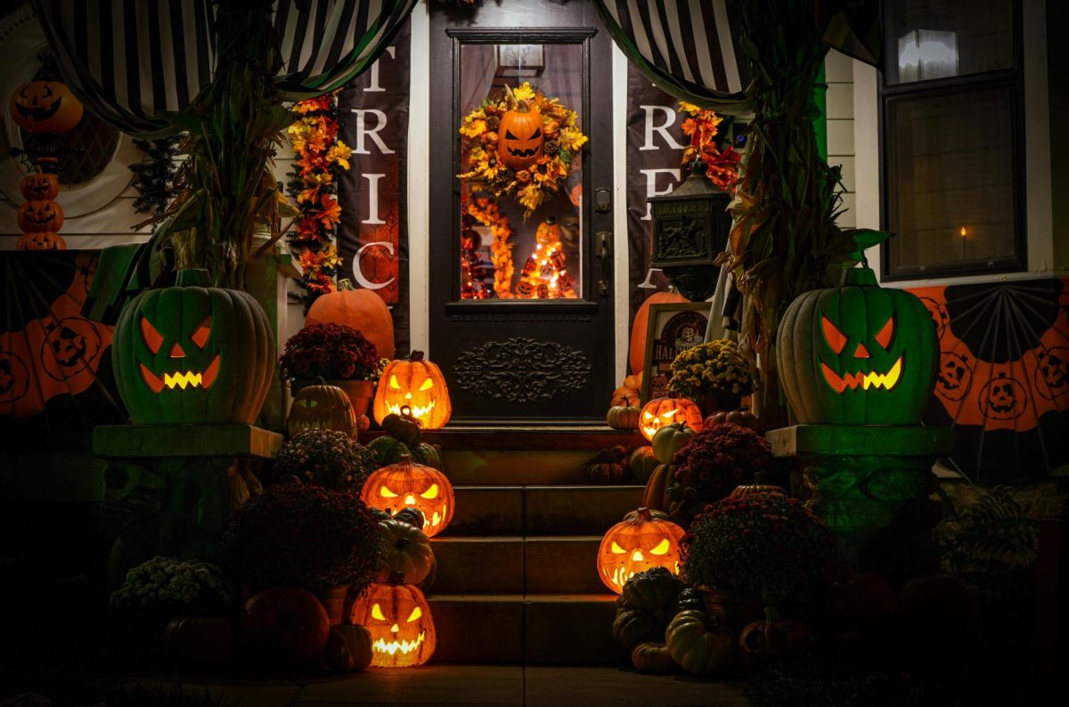 A front porch is decorated with lit orange jack-o-lanterns up and down the steps, gourds, and flowers. An eerie pumpkin face is hung on the door with a fall wreath around it.