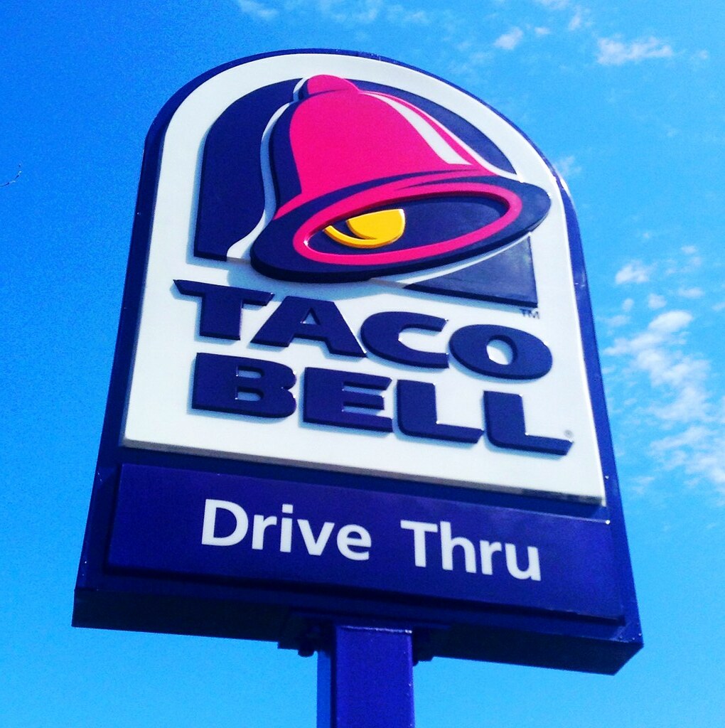 The image shows a modern style Taco Bell sign with the "Drive Thru" lettering at the bottom of the blue, pink and yellow themed logo.