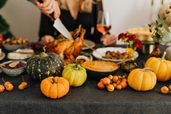 The table features various gourds in the foreground and Thanksgiving foods in the background including cranberry relish, squash, stuffing, pies and a wine glass. Someone is carving a turkey in the back center.