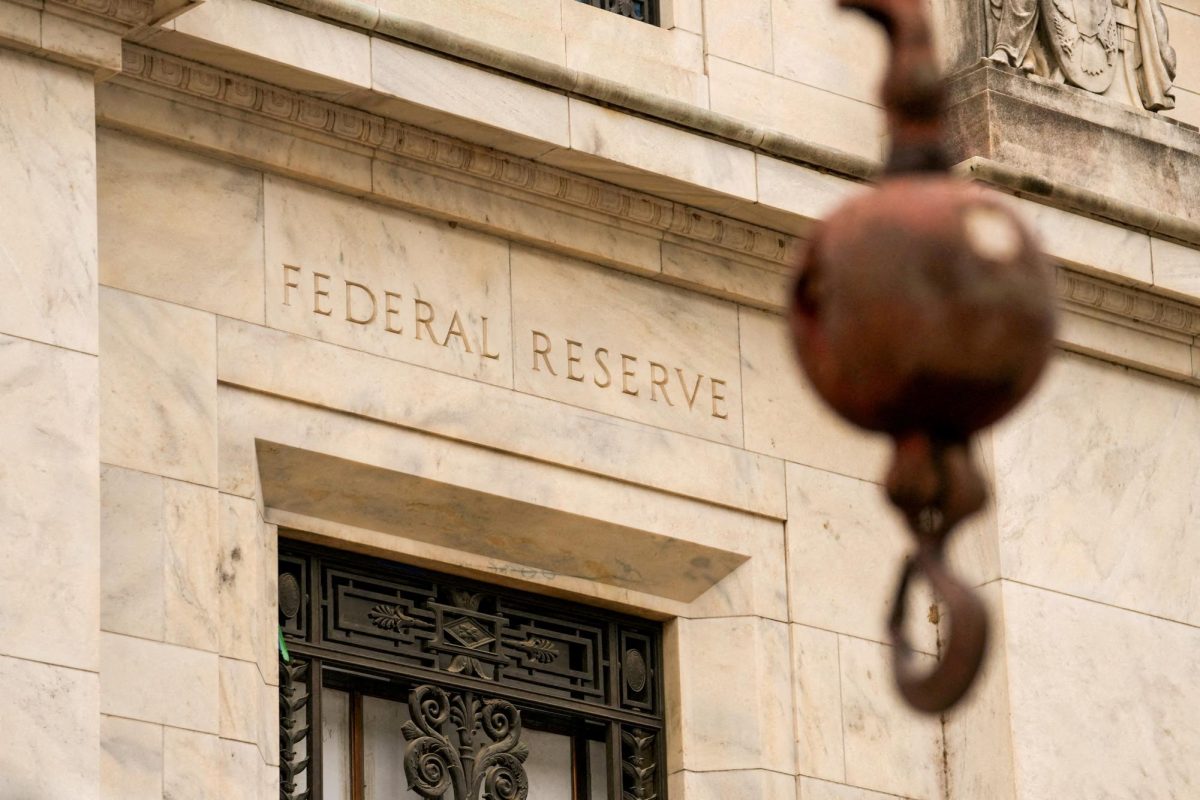 The image shows the exterior of the Federal Reserve Board building with sand colored stone, an ornamental wrought iron door, and a copper street unfocused in the foreground.