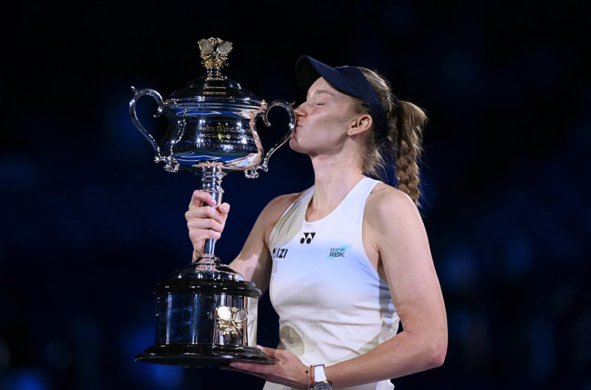 Rybakina, dressed in white with a navy hat, kisses the championship trophy.