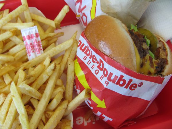 The image shows a red/white/yellow packaged burger which is stuffed with bacon, cheese, lettuce, pickle. A side of fries is placed next to the burger.