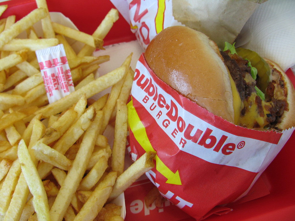 The image shows a red/white/yellow packaged burger which is stuffed with bacon, cheese, lettuce, pickle. A side of fries is placed next to the burger.