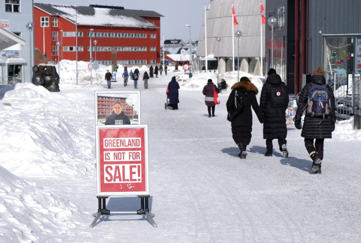 People walk on a snow covered road. The prominent sign says "Greenland is not for sale!" in red and white.
