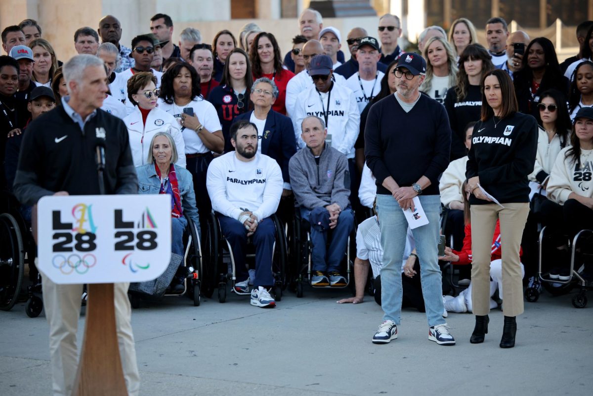 The speaker stands in the foreground, speaking in a microphone at a podium that says LA 28 on it with the olympics and paralympics logos.