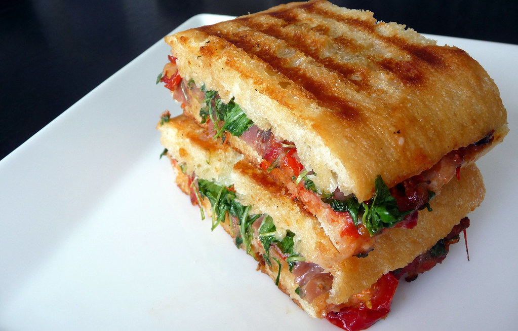 A panini consisting of cured meats, cheese, tomatoes and spinach on focaccia bread rests on a white plate