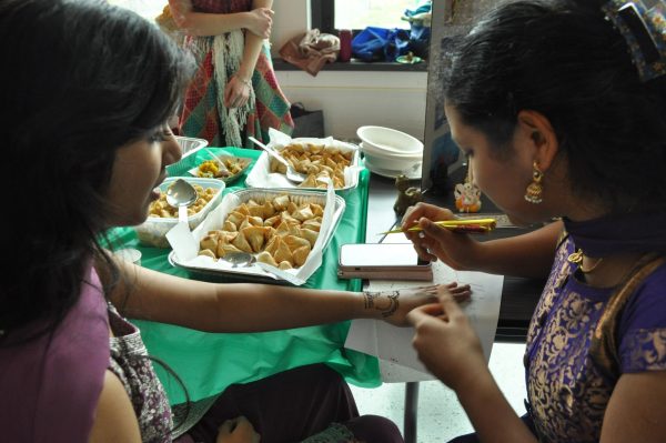 A student draws Henna art on another students hand. Traditional food is on the table in the background.