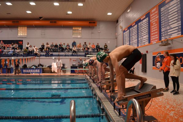 The swimmers are crouched down, holding the handles on the platforms. An FM swimmer is in the closest lane with a green and white cap on.
