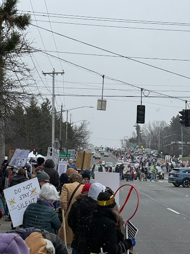 The shot looks up the sloped road where people are gathered on both sides of the road with signs. Cars are stopped by protesters in the road.