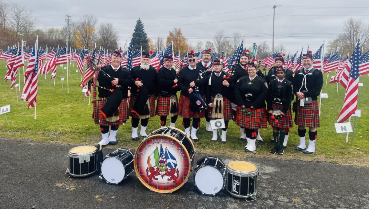 Members of the band pose in their red and black kilts and black jackets in front of a field filled with American flags.