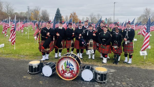Members of the band pose in their red and black kilts and black jackets in front of a field filled with American flags.